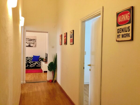A hallway with a wooden floor, a white door, and a wall with a 'WARNING GENIUS AT WORK' sign. There is a couch with a blue pillow and a plant in the background.