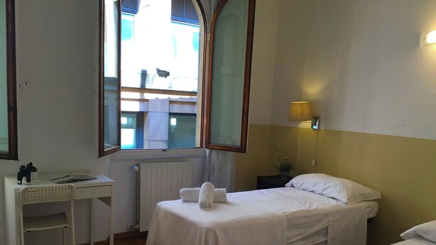 Quarto de hotel com duas camas de solteiro, uma escrivaninha, uma cadeira, uma janela com vista para o exterior e uma luminária fixada na parede.