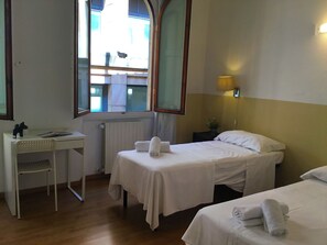 A hotel room with two single beds, a desk, a chair, a window with a view of the outside, and a wall-mounted lamp.