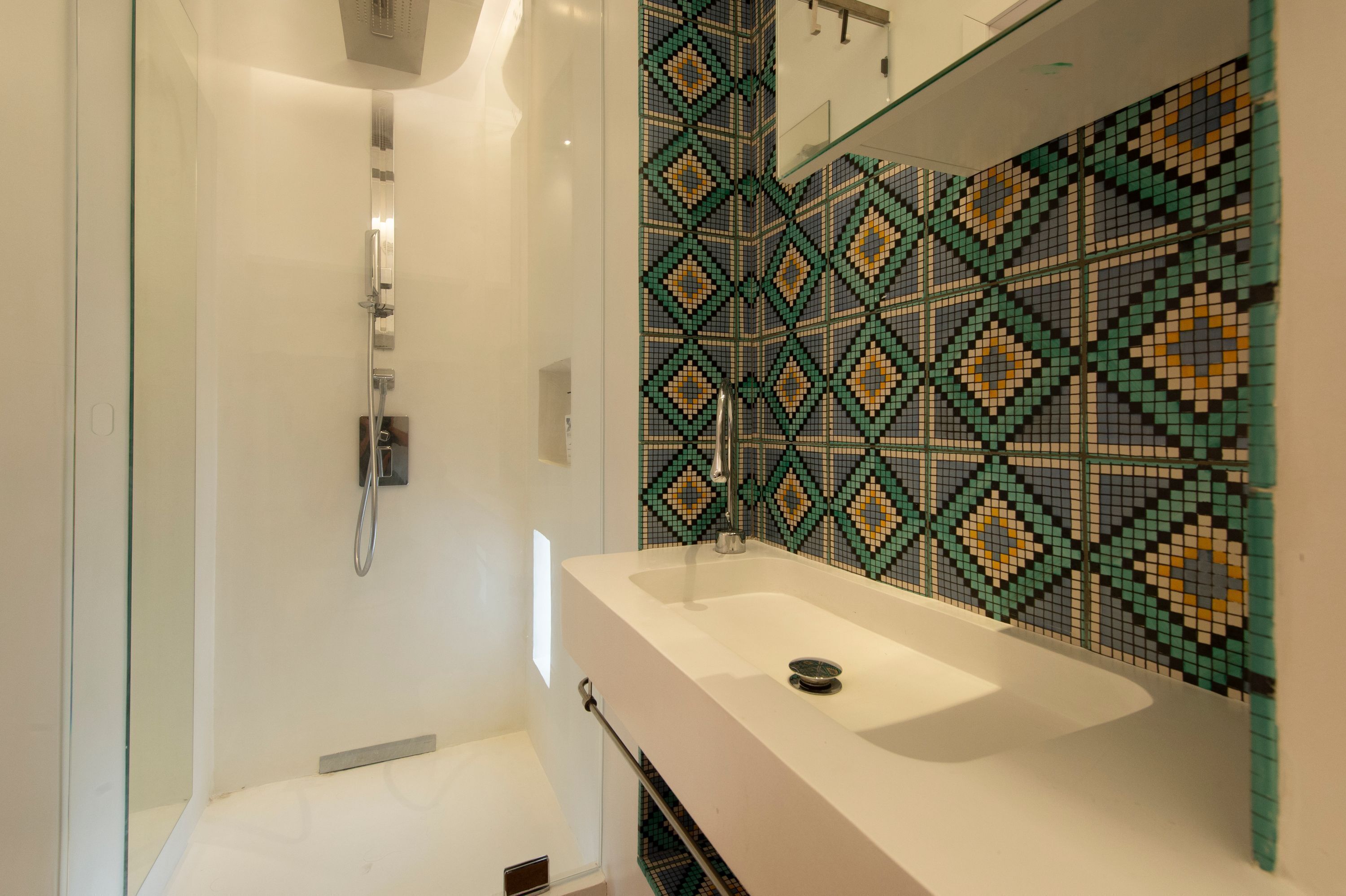 Un baño con un lavamanos blanco, cabina de ducha con mampara de vidrio y pared revestida con un patrón geométrico.