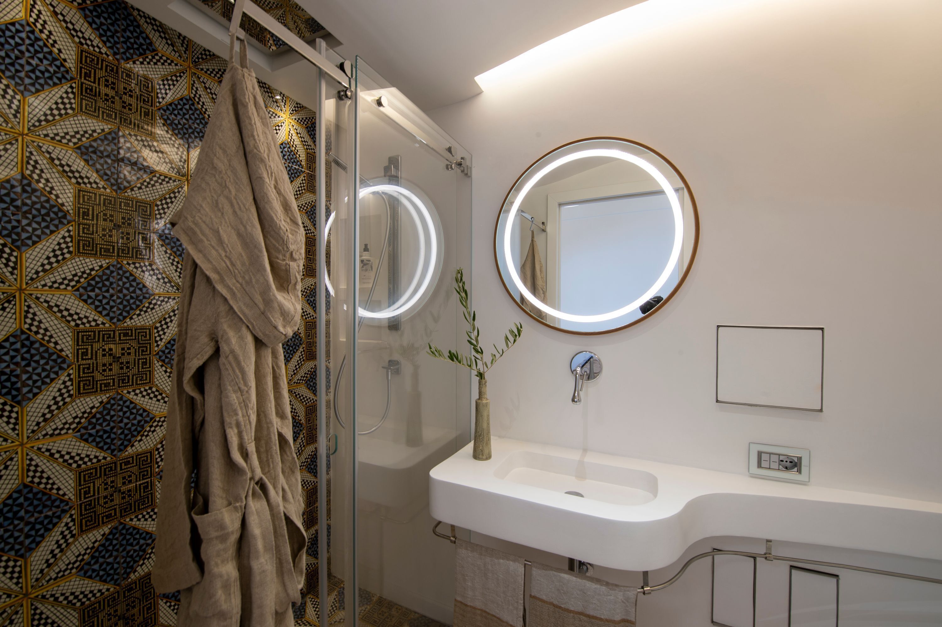 Ein Badezimmer mit Duschkabine aus Glas, einem weißen Waschbecken mit rundem Spiegel und einer Wand mit Musterung.