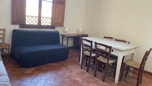Une pièce avec un canapé bleu, une table de salle à manger en bois et quatre chaises, une petite table d’appoint et une fenêtre avec des volets en bois.