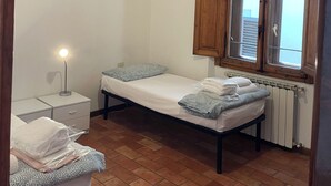 Une petite chambre propre, avec deux lits simples, une table de chevet, une lampe et une fenêtre avec des volets en bois.