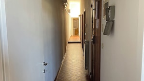 Un couloir étroit, dont le sol est recouvert d’un parquet à motifs en chevrons, avec des murs blancs et une porte au fond qui donne sur une pièce lumineuse.