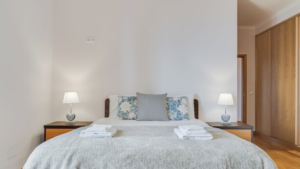 A neatly made bed with a grey blanket, two bedside tables with lamps, and a wooden wardrobe in a well-lit room.