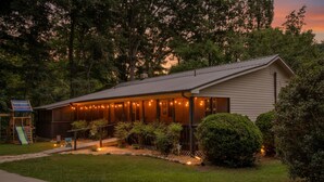 A two-story building with a porch, string lights, and a small play area. - Blue Ridge Haven is packed with luxury and family fun including a Hot Tub, Creekside Fire Pit, Game Room, Private Yard with Playset, and Back Deck with Grill. Your stay is owned and managed by a four time AirBnB Superhost. (Blue Ridge)