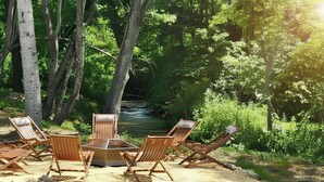 A forested area with a river, surrounded by trees and a fire pit with chairs. - Blue Ridge Haven is packed with luxury and family fun including a Hot Tub, Creekside Fire Pit, Game Room, Private Yard with Playset, and Back Deck with Grill. Your stay is owned and managed by a four time AirBnB Superhost. (Blue Ridge)
