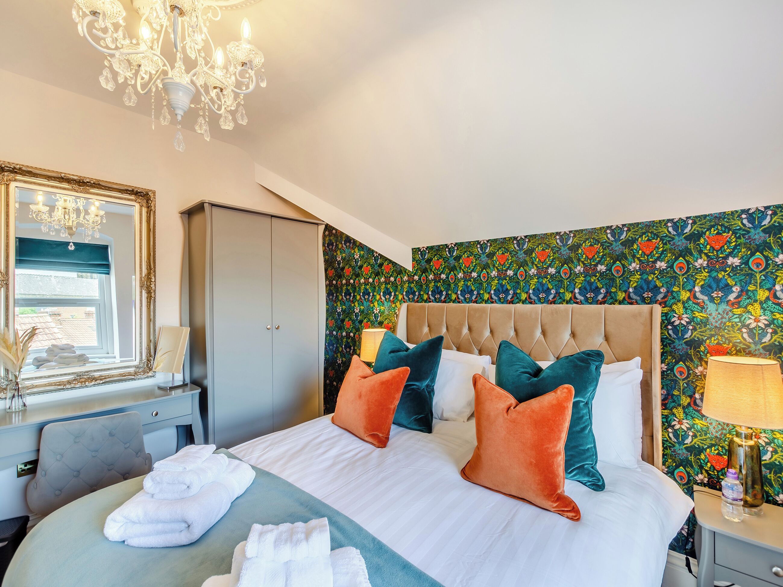 A hotel room with a bed, a chandelier, a wardrobe, and a colorful floral wallpaper.
