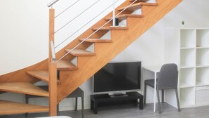 A modern staircase with wooden steps and metal railings, a flat-screen TV on a black stand, and a grey chair in a minimalistic room. - Stunning, Bright Duplex (Saint-Ouen-sur-Seine)