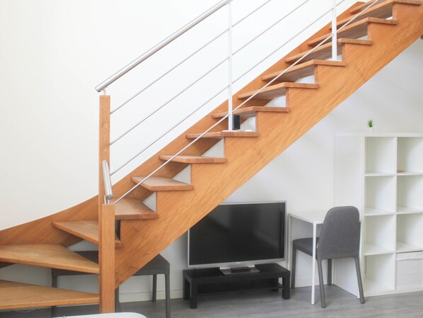 Un escalier moderne avec des marches en bois et des rampes en métal, un téléviseur à écran plat posé sur un meuble noir, et un fauteuil gris dans une pièce au décor minimaliste.