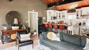 A modern living room with a grey sofa, a black leather chair, and a wooden coffee table. A kitchen with white cabinets and red bar stools is visible in the background. - Fully Remodeled Home Away From Home with all the amenities (Sugarloaf)