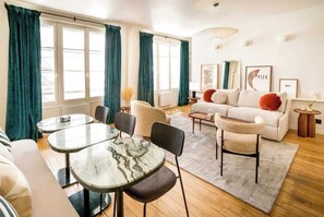 Uma sala de estar moderna com um sofá, duas poltronas, uma mesa de centro e uma mesa de jantar. Há cortinas nas janelas, um abajur e quadros na parede.