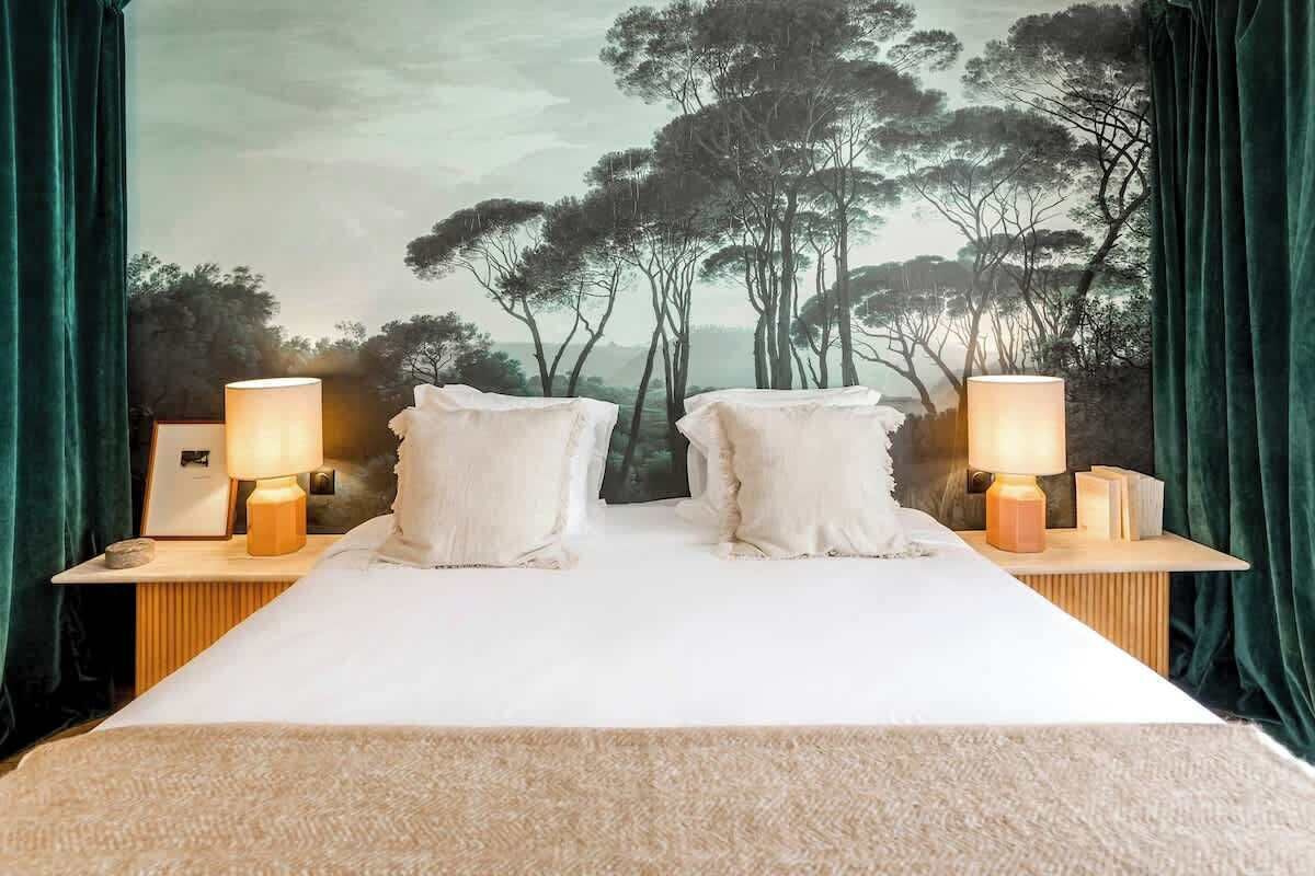 Een bed met twee kussens, twee nachtkastjes met lampen, en een muurschildering van bomen.