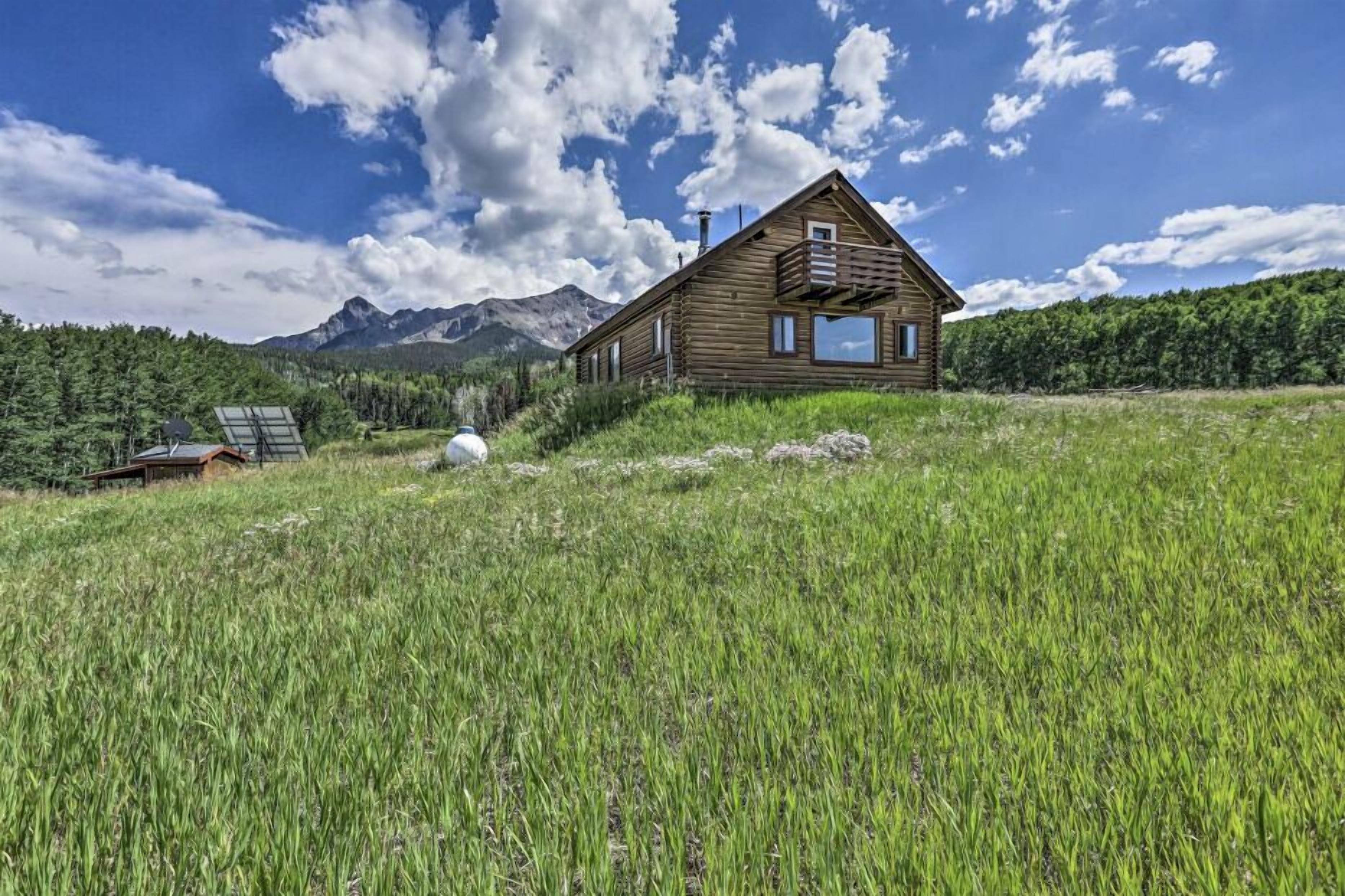 A wooden cabin with a balcony, surrounded by a grassy field and mountains in the background.