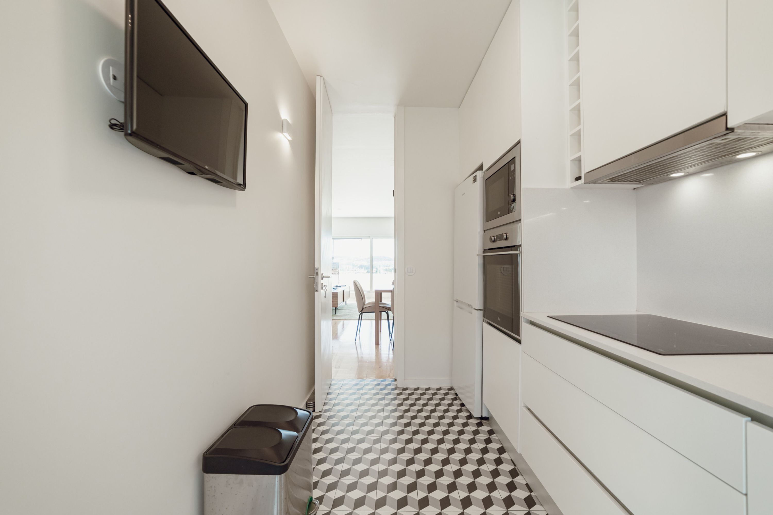 A narrow hallway with a tiled floor, a trash bin, a flat-screen TV mounted on the wall, and a kitchen area with a stove and oven.