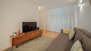 A modern living room with a flat-screen TV on a wooden console, a brown sofa with cushions, and a large window with sheer curtains. - Porto Panoramic Views With Terrace by Vacationy (Porto)