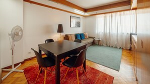 Um espaço compacto para viver, com uma mesa de jantar, cadeiras, um sofá, uma pequena mesa lateral, um ventilador e uma janela com cortinas.
