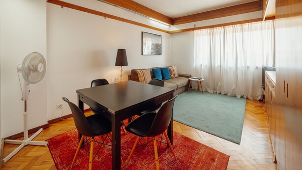 Un espace de vie compact comprenant une table à manger, des chaises, un canapé, une petite table d’appoint, un ventilateur et une fenêtre avec des rideaux.