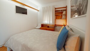 Quarto com uma cama, escrivaninha, cadeira, televisão e janela com cortinas.