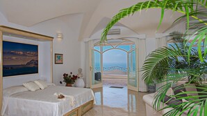 Una camera da letto con un letto, un quadro, una pianta in vaso e vista sul mare attraverso una porta aperta.