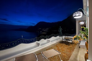Un balcón con vista al mar y a las montañas, una mesa con sillas e iluminación decorativa.