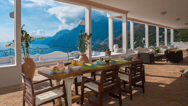Zona pranzo con tavolo apparecchiato per quattro, ampie finestre che offrono vista su montagne e mare, e un balcone con posti a sedere esterni.