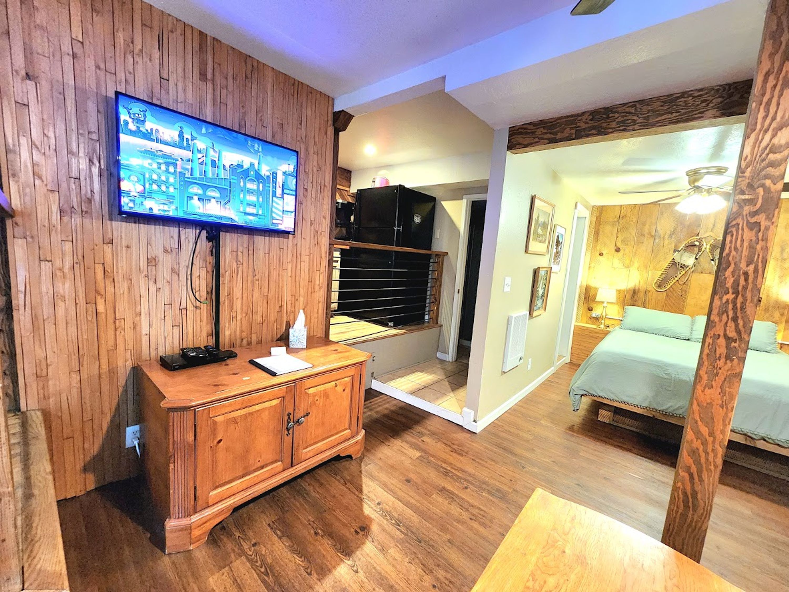 A hotel room with a wooden wall, a television mounted above a wooden cabinet, a bed with a headboard, and a ceiling fan.