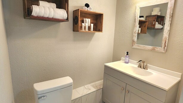 Un baño con inodoro, lavabo con espejo y dos repisas con artículos de aseo.