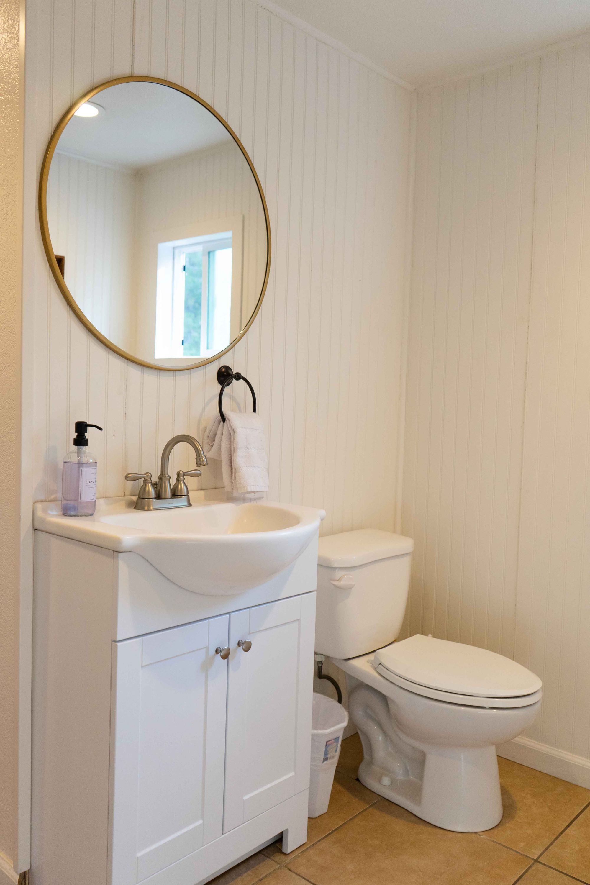 A bathroom with a white vanity, a round mirror, a toilet, and a window.