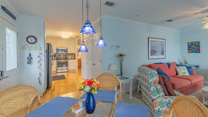 A well-lit living room with a sofa, dining table, and chairs. There is a kitchen in the background with cabinets and a refrigerator. - Bear Point Bayview Cottage – Bay Views, Marina Walk (Orange Beach)