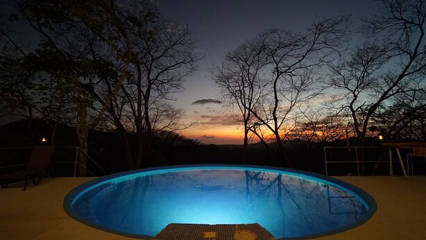 Une piscine au crépuscule, avec vue sur le coucher de soleil, entourée d’arbres.