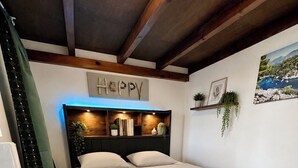 A bedroom with a bed, wooden headboard, shelves with books and plants, a 'happy' sign, and a framed picture on the wall. - Appartement Cosy - Chartreux (Marseille)