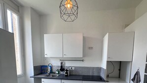 A compact kitchen with white cabinets, a black countertop, a hanging light fixture, and a chair with a patterned tablecloth. - Appartement Cosy - Chartreux (Marseille)