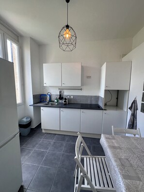 A compact kitchen with white cabinets, a black countertop, a hanging light fixture, and a chair with a patterned tablecloth.