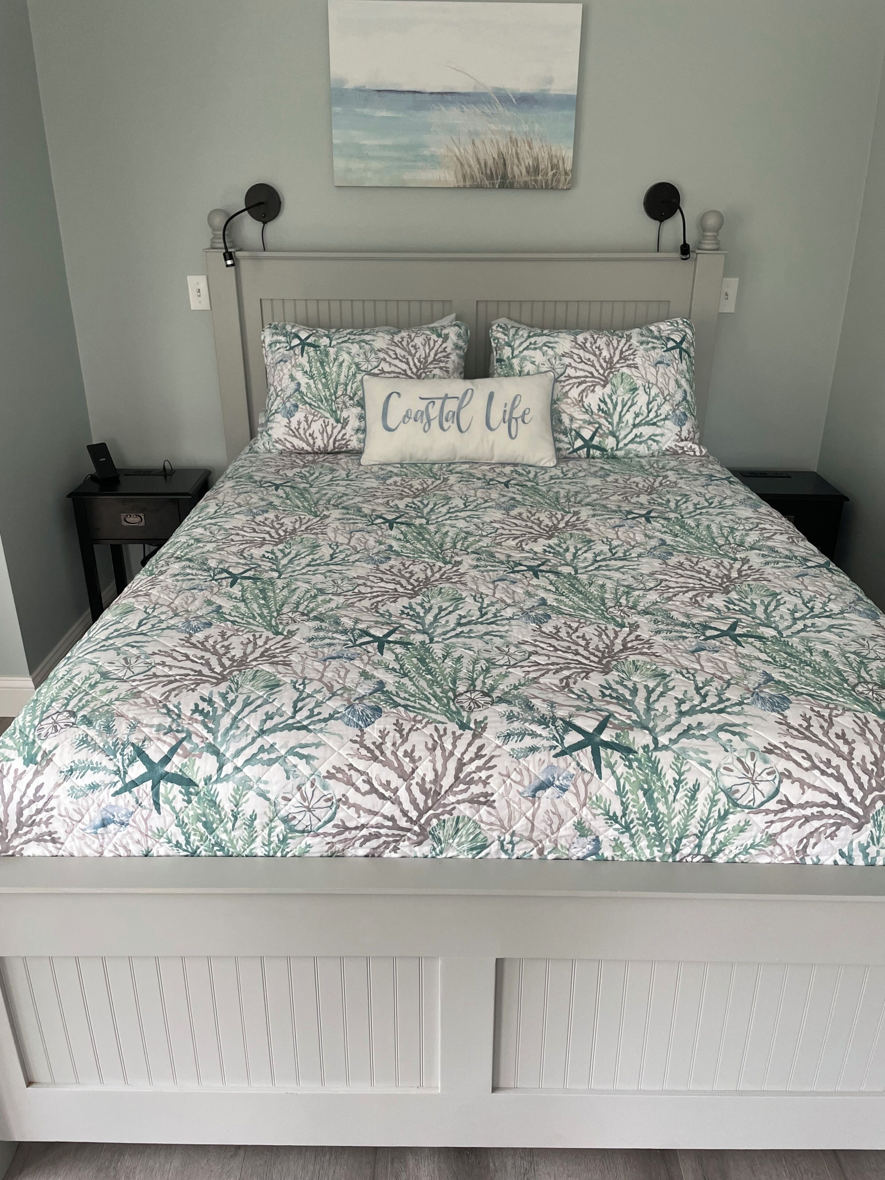 A bed with a coastal life themed bedspread, a bedside table with a phone, and a wall-mounted lamp.