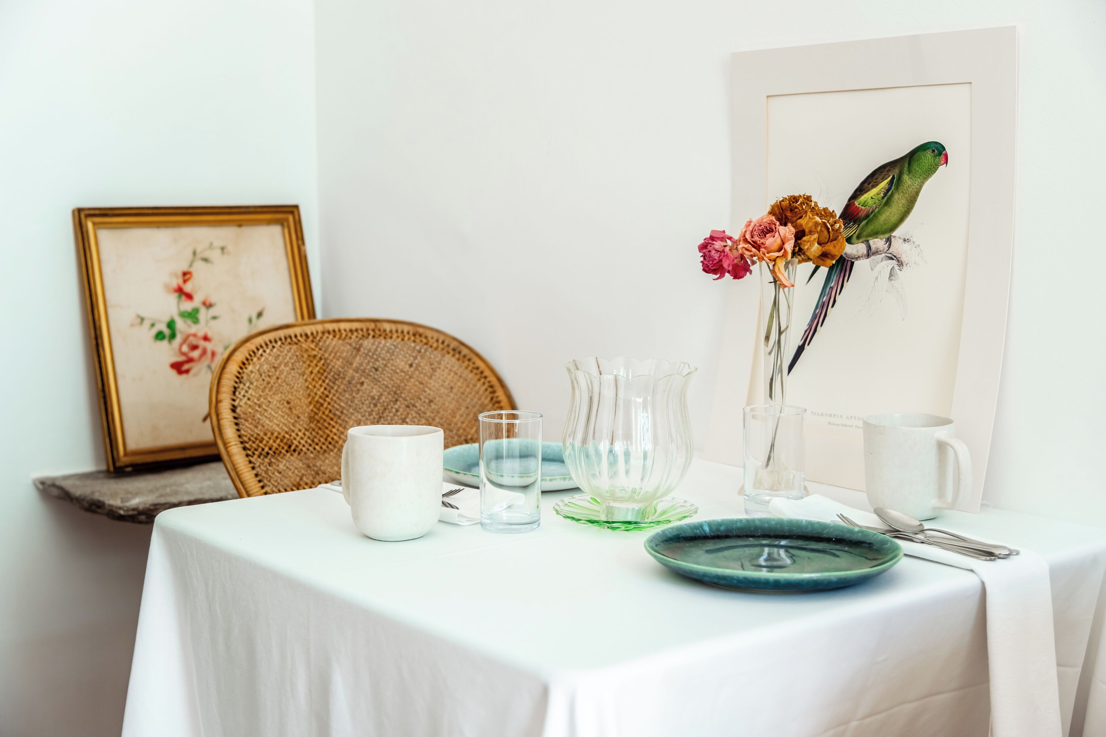 A dining table with a white tablecloth, a wicker chair, a framed floral artwork, a vase with flowers, and a parrot illustration.
