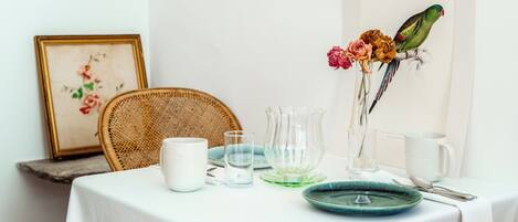 A dining table with a white tablecloth, a wicker chair, a framed floral artwork, a vase with flowers, and a parrot illustration.