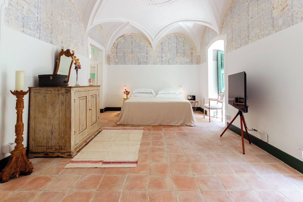 A bedroom with a bed, a dresser, a television on a tripod, and a large window.