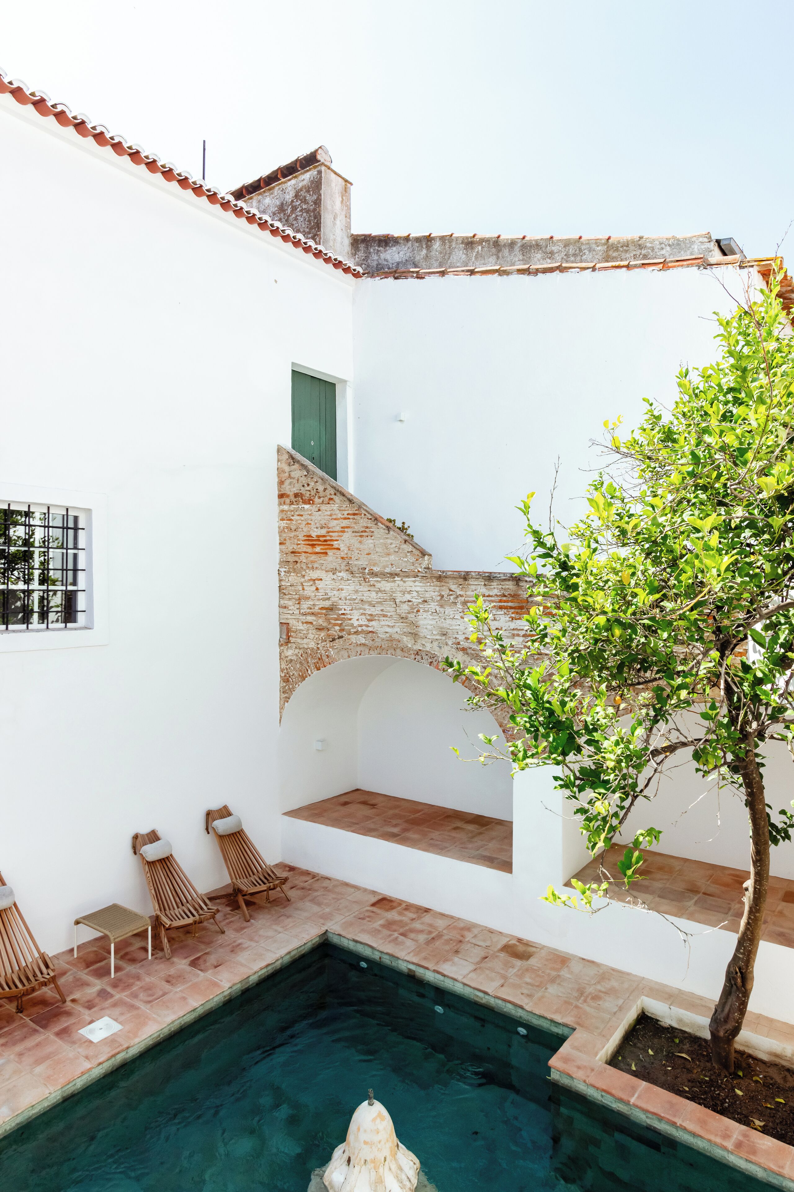 A pool area with a clear pool, wooden lounge chairs, a small table, and a tree.
