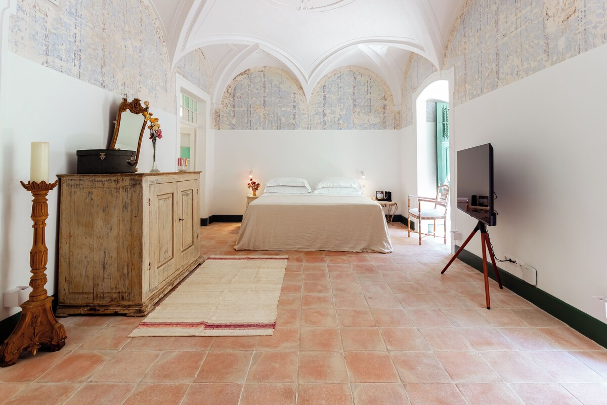 A bedroom with a bed, a dresser, a television on a tripod, and a large window.