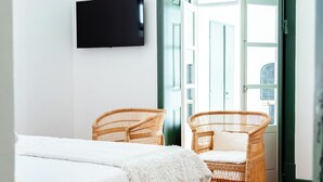 Ett sovrum med en säng, två fåtöljer i rotting, en tv på väggen och en grön dörr som leder ut.