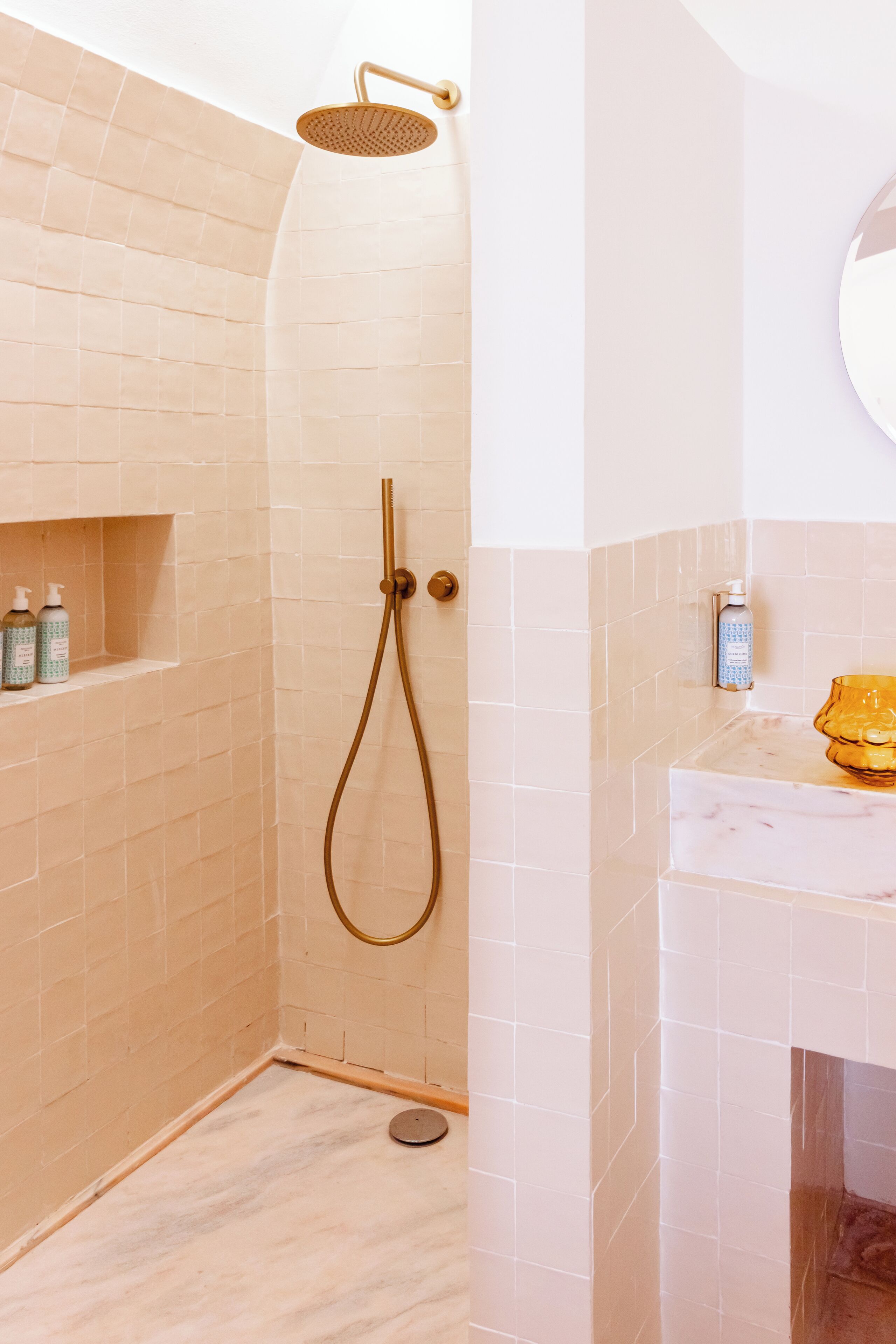 Ein Badezimmer mit beigelfarben gefliesten Wänden, einer Dusche mit goldfarbener Duschbrause und einer Marmorplatte mit Toilettenartikeln.