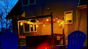 Un foyer extérieur devant une maison, avec deux chaises bleues et des guirlandes lumineuses.