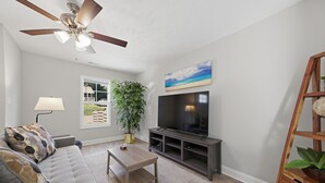 Ein Wohnzimmer mit grauem Sofa, einem Couchtisch, einem Fernseher auf einem Holzständer, einem Deckenventilator, einem Fenster mit Aussicht und einer Pflanze.