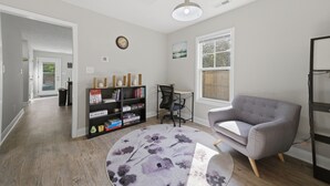 Ein Wohnzimmer mit grauem Sofa, einem runden Teppich, einem Bücherregal und einem Schreibtisch.