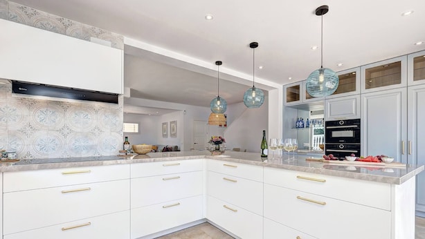 Une cuisine moderne avec des meubles blancs, des poignées dorées et un plan de travail en marbre. On y trouve des luminaires suspendus et un four encastré.