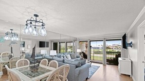 A spacious living room with a grey sofa, wicker chairs, and a glass dining table. There is a large window offering a view of the outdoors. - Fall Deals + Snowbird Rates | Coastal Stay Near the Beach (Panama City Beach)