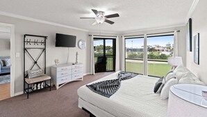 A bedroom with a large bed, a ceiling fan, a TV, a desk, and a view of the outdoors. - Fall Deals + Snowbird Rates | Coastal Stay Near the Beach (Panama City Beach)