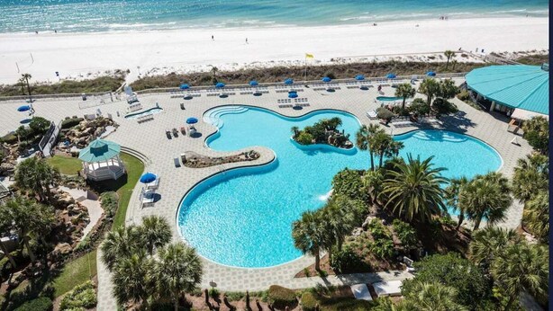 A large, irregularly shaped swimming pool with a beach view, surrounded by lounge chairs and umbrellas. - Fall Deals + Snowbird Rates | Coastal Stay Near the Beach (Panama City Beach)