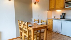 A dining area with a wooden table and chairs, a kitchen with wooden cabinets and a white dishwasher, and a wall-mounted lamp. - 40m from ski slopes, floor -2, fitness, swimming pool, sauna, balcony, tv, ski locker, 31m² (Les Orres)